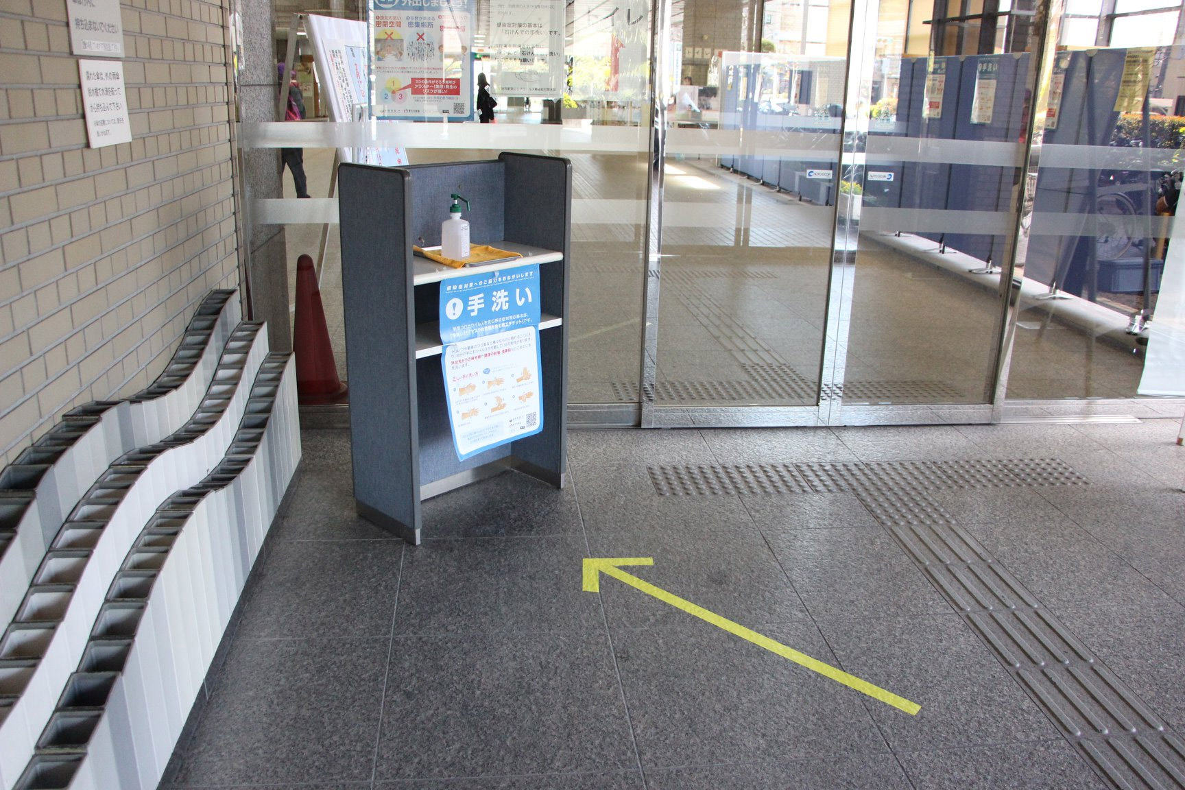 写真)京都府宇治市 庁舎1階正面玄関をはじめ、出入口の複数個所に、数歩手前から消毒液へ誘導する矢印を示したイエロ-テープを貼り付け、消毒液の使用を促している。