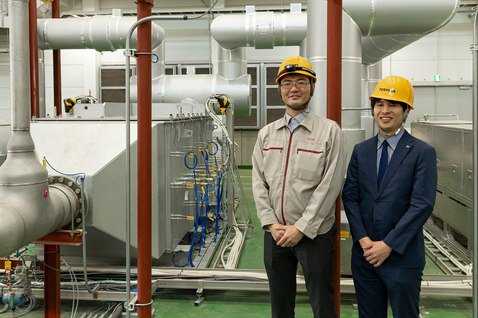 写真)岩石蓄熱試験設備の蓄熱槽の前にて 東芝エネルギーシステムズ株式会社パワーシステム事業部新規事業開発部 三木 浩睦氏(右)、同エネルギーシステム技術開発センター機械技術開発部 馬渡 峻史氏